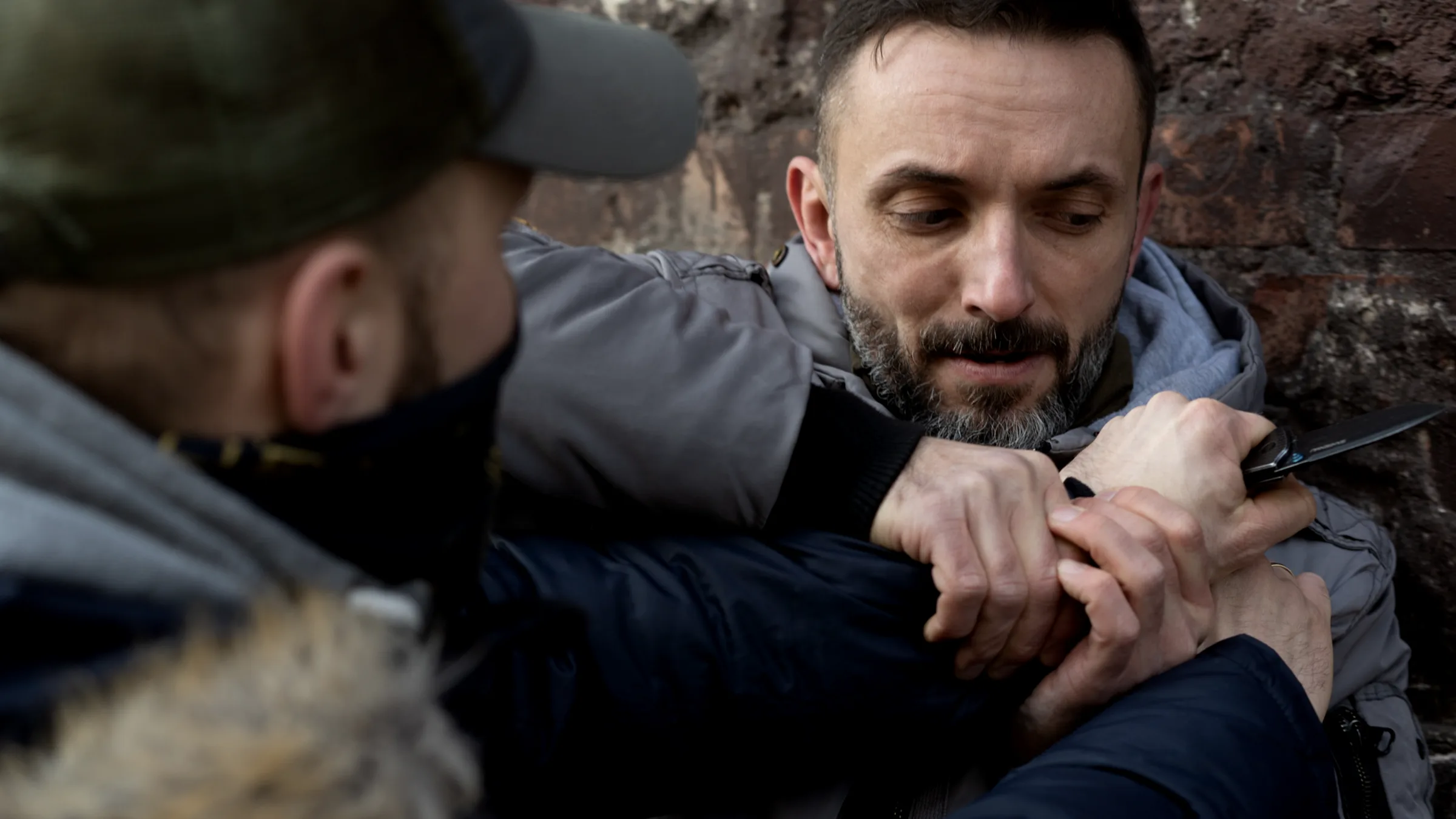 A knife-defence drill close-up — both hands pinning the attacker's blade hand against the body